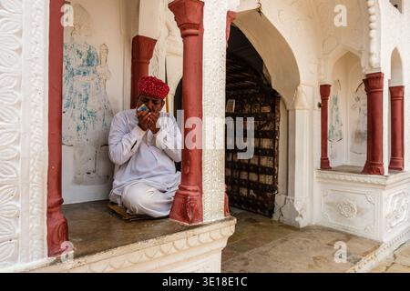 Un homme dans un turban rouge est assis dans une alcôve blanche à Pushkar, allumant une cigarette tandis qu'une lourde porte en bois est vue au loin. Banque D'Images