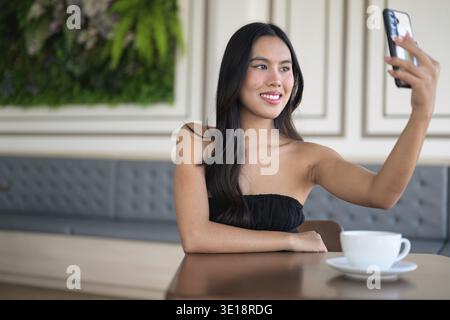 Belle jeune femme asiatique assise dans un café à l'aide d'un téléphone portable tout en ayant une tasse à café sur la table pendant le concept de travail à distance de jour Banque D'Images