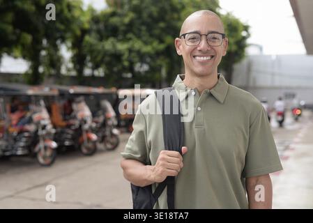 Homme chauve latin en polo décontracté et sac à dos debout à l'extérieur en Thaïlande avec des taxis tuk tuk derrière lui. Voyager, visiter et explorer ainsi Banque D'Images