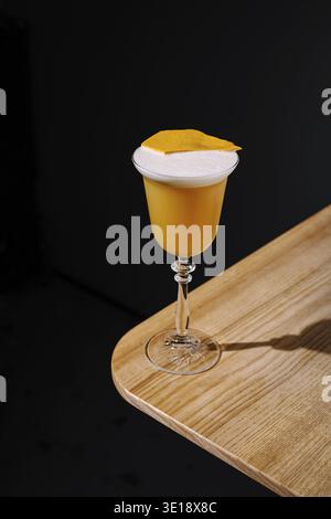 Un cocktail vibrant se tient élégamment sur une table en bois, avec une boisson jaune onctueuse surmontée d'une couche mousseuse et d'une garniture d'agrumes. Le backgro sombre Banque D'Images