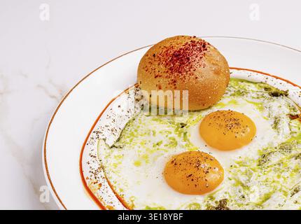 Une assiette présente trois œufs ensoleillés recouverts d'une sauce verte éclatante et d'une pincée d'épices, accompagnés d'un petit pain moelleux avec une saison rouge Banque D'Images
