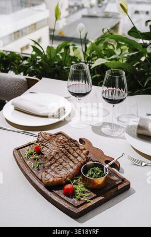 Un dîner de steak magnifiquement arrangé comprend une coupe grillée sur une planche de bois, accompagnée de riches herbes vertes et tomates cerises. Deux verres de rouge Banque D'Images