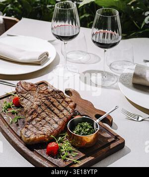 Un steak grillé magnifiquement présenté se trouve sur un plateau en bois à côté d'herbes fraîches. Deux élégants verres de vin rouge vous attendent dans un élégant dîner en plein air Banque D'Images