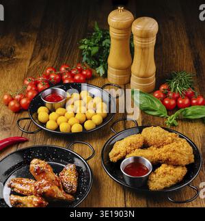 Une variété de hors-d'œuvre, y compris des ailes de poulet croustillantes, des boulettes de fromage doré et des collations frites, sont présentés sur des assiettes sombres. Tomates fraîches et herbes e Banque D'Images