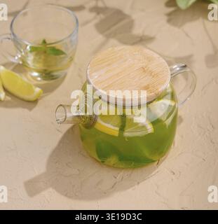 Théière en verre ensoleillée avec thé à la menthe rafraîchissant et citron sur fond beige Banque D'Images