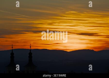 Des clochers d'église silhouettés se dressent devant un ciel orange et jaune spectaculaire, superposé sur des montagnes lointaines au crépuscule. Banque D'Images