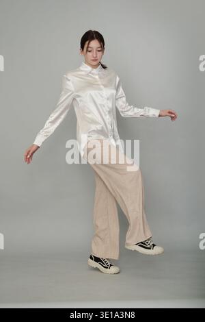 Une jeune femme dans une blouse blanche satinée et un pantalon beige pose gracieusement sur une jambe sur un fond neutre. Cette image souligne l'élégance et Banque D'Images