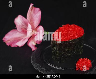 Sushi Gunkan maki avec tobiko de caviar rouge décoré de fleurs roses sur fond noir Banque D'Images