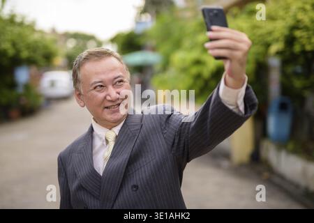 Portrait d'un homme d'affaires asiatique mûr portant le costume tout en prenant selfie avec téléphone mobile dans les rues à l'extérieur Banque D'Images