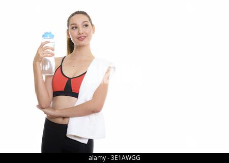 Studio shot of young happy Asian woman smiling et penser tout en tenant la bouteille de l'eau prêt pour salle de sport Banque D'Images