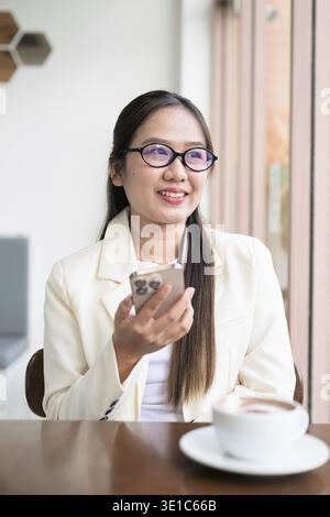 Jeune femme d'affaires asiatique assise dans un restaurant de café portant costume et lunettes tout en utilisant un smartphone Banque D'Images