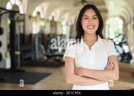 Belle femme asiatique en polo blanc posant à l'intérieur de la salle de gym. Concept de style de vie de fitness élégant avec un équipement moderne en arrière-plan. Détendu et sportif Banque D'Images