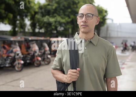 Homme chauve latin en polo décontracté et sac à dos debout à l'extérieur en Thaïlande avec des taxis tuk tuk derrière lui. Voyager, visiter et explorer ainsi Banque D'Images