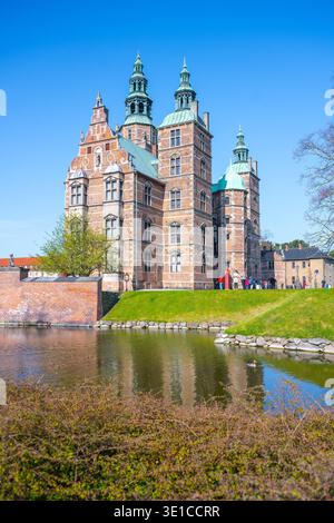 Les visiteurs admirent l'architecture étonnante du château de Rosenborg reflétée dans les eaux tranquilles des douves environnantes. Le ciel bleu vif rehausse la beauté de ce site historique. Banque D'Images