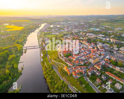 Cette vue aérienne à couper le souffle capture la ville de Melnik et son château historique surplombant la paisible rivière Labe en Tchéquie. Le paysage présente une végétation luxuriante et une architecture charmante pendant le coucher du soleil. Banque D'Images