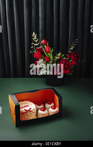 Une boîte magnifiquement agencée présente des cupcakes décorés avec des garnitures de coeur à côté d'un bouquet vibrant de fleurs rouges. La toile de fond est sombre, ele Banque D'Images