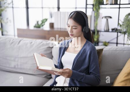 Concentré jeune femme asiatique recherche étudiante avec livre et écouteurs sur canapé à la maison étudier en ligne Banque D'Images