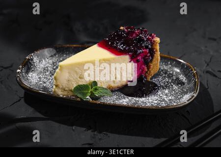 Tranche de cheesecake crémeux garnie d'une compote de baies vibrante et de sucre en poudre, élégamment servie sur une assiette sombre Banque D'Images