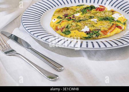Omelette aux légumes et fromage de chèvre servie sur une assiette avec couverts sur une table avec une nappe blanche Banque D'Images