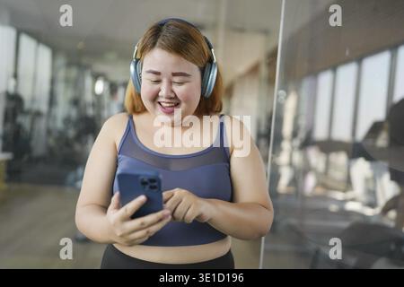 Femme avec téléphone intelligent après l'entraînement à Gym. Femme asiatique obèse regardant la classe de formation d'entraînement en ligne Banque D'Images