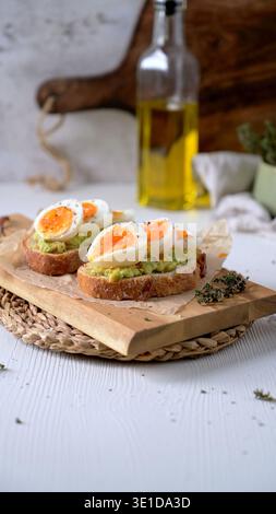 Deux tranches de pain au levain croustillant avec de la purée d'avocat et des œufs à la coque sur une planche de bois. Un éclairage naturel lumineux met en valeur la texture crémeuse Banque D'Images