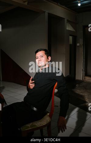 Un homme dans une chemise noire à manches longues est assis sur une chaise, regardant directement le spectateur. Le cadre intérieur offre un éclairage spectaculaire et des ombres profondes, cr Banque D'Images