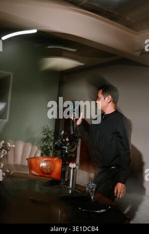Un homme concentré dans une chemise sombre vérifie soigneusement son appareil photo monté sur un stabilisateur de cardan professionnel à l'intérieur. Il se prépare pour un tournage, assurant un Banque D'Images