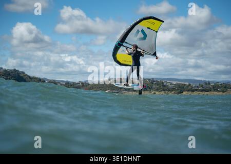 Une aile d'homme défile en mer à l'aide d'une aile gonflable tenue à la main, chevauchant une planche de surf hydrofoil à Plimmerton Beach, NZ. Mer turquoise, ciel bleu nuageux. Banque D'Images