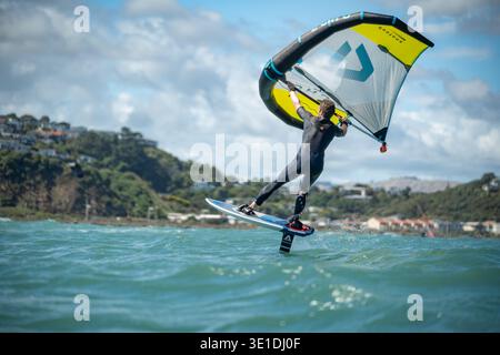 Une aile d'homme défile en mer à l'aide d'une aile gonflable tenue à la main, chevauchant une planche de surf hydrofoil à Plimmerton Beach, NZ. Mer turquoise, ciel bleu nuageux. Banque D'Images