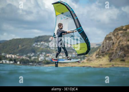 Une aile d'homme défile en mer à l'aide d'une aile gonflable tenue à la main, chevauchant une planche de surf hydrofoil à Plimmerton Beach, NZ. Mer turquoise, ciel bleu nuageux. Banque D'Images