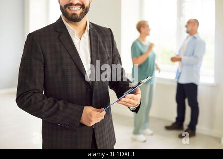 Agent immobilier souriant avec presse-papiers rencontre couple dans la maison lumineuse ensemble Banque D'Images