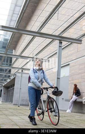 Femme dans la vingtaine marchant et tenant un vélo de route avec casque sous la verrière, portant une veste en denim Banque D'Images