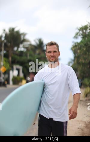 Scène côtière avec surfer occasionnel. Jeune homme décontracté se penchant sur la planche de surf au milieu de la côte Banque D'Images