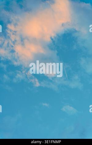 Ciel bleu éclatant avec des nuages rose-orange moelleux et une traînée blanche d'avion qui se répand à travers, capturant la beauté atmosphérique sereine au coucher du soleil. Banque D'Images