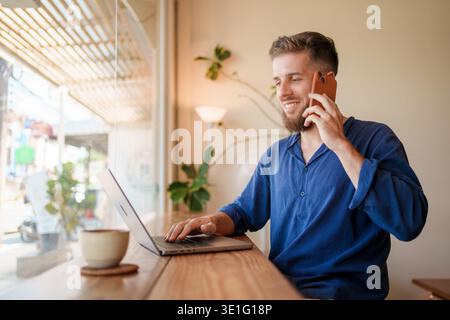 Un jeune homme travaille sur son ordinateur portable et son smartphone dans un café confortable, profitant d'une journée productive Banque D'Images