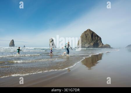 Mère, fils et fille courent des vagues hors de l'océan devant Haystack Rock sur Cannon Beach, Oregon. Banque D'Images