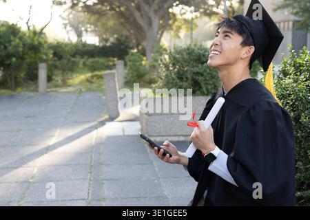 Homme asiatique assis sur le chemin du campus tenant le diplôme et le smartphone dans la robe et mortarboard, espace de copie Banque D'Images