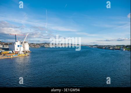 L'infrastructure portuaire industrielle avec de grandes grues de construction surplombe les eaux bleues et la côte résidentielle de Haugesund, en Norvège dans cette scène maritime et de voyage. Banque D'Images