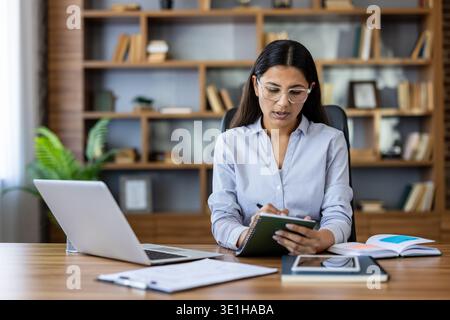Focalisé jeune femme d'affaires dans un bureau moderne, prendre des notes tout en multitâche sur un ordinateur portable et organiser la paperasse pour la stratégie, la planification et le développement de carrière Banque D'Images