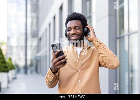 Jeune homme souriant dans une rue urbaine, écoutant de la musique et des podcasts sur des écouteurs sans fil tout en interagissant avec son smartphone pendant un trajet quotidien détendu Banque D'Images