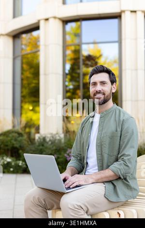 Homme souriant dans des vêtements décontractés intelligents travaillant à distance sur un ordinateur portable, assis sur un banc en bois dans un cadre de parc urbain moderne par un bâtiment avec des fenêtres réfléchissantes, embrassant le travail flexible et la technologie Banque D'Images