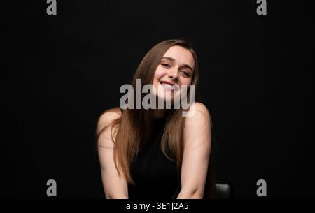 Jeune femme avec de longs cheveux bruns souriant à la caméra, sentant le bonheur et regardant confiant sur fond noir Banque D'Images