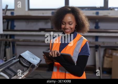 Portrait d'une travailleuse afro-américaine souriante dans un gilet orange de sécurité utilisant une tablette numérique dans une usine de traitement des métaux. Banque D'Images