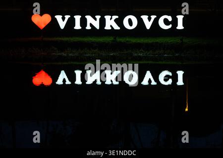 VINKOVCI, CROATIE - février 18.2026:grand cœur incandescent et lettres Vinkovci allumées la nuit sur une rive, reflétées dans l'eau calme. Atmosphérique urbaine Banque D'Images