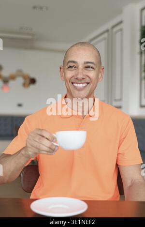 Bel homme chauve italien d'âge moyen souriant dans un polo orange assis à la table de café tenant une tasse de café. Concept de style de vie décontracté Banque D'Images
