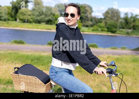 Femme faisant du vélo le long de la rivière chemin parc ensoleillé, profiter de l'activité de plein air de l'air frais. Style de vie détendu liberté de scène loisirs mouvement sain dans Banque D'Images