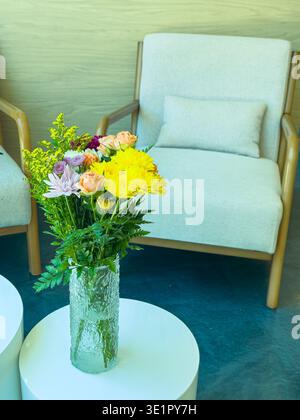 Un vase en verre contient une variété de fleurs, placées sur une table blanche à côté d'une chaise de couleur claire dans un espace intérieur bien éclairé. La lumière du soleil remplit la zone, hig Banque D'Images