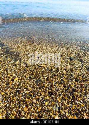 De petits cailloux et du sable sont visibles sur la plage avec de l'eau se déplaçant doucement sur la surface. Le soleil brille brillamment dans le ciel pendant les heures de lumière du jour. Banque D'Images