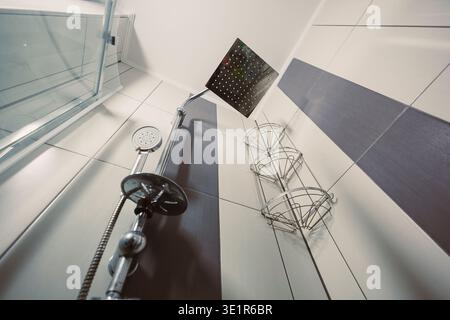 L'eau coule de la pomme de douche dans une salle de bains moderne. Les étagères de rangement peuvent contenir des articles de bain. La lumière du soleil éclatante entre par une fenêtre voisine. Les murs le sont Banque D'Images