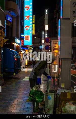 Un travailleur sanitaire de fin de nuit portant des gants bleus et un couvre-tête ramasse les ordures dans une rue éclairée au néon dans le quartier de Roppongi à Tokyo, au Japon. Banque D'Images